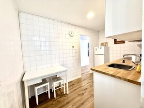 A compact kitchen with white tiled walls, a wooden countertop, a sink, a refrigerator, and a small dining table with chairs.