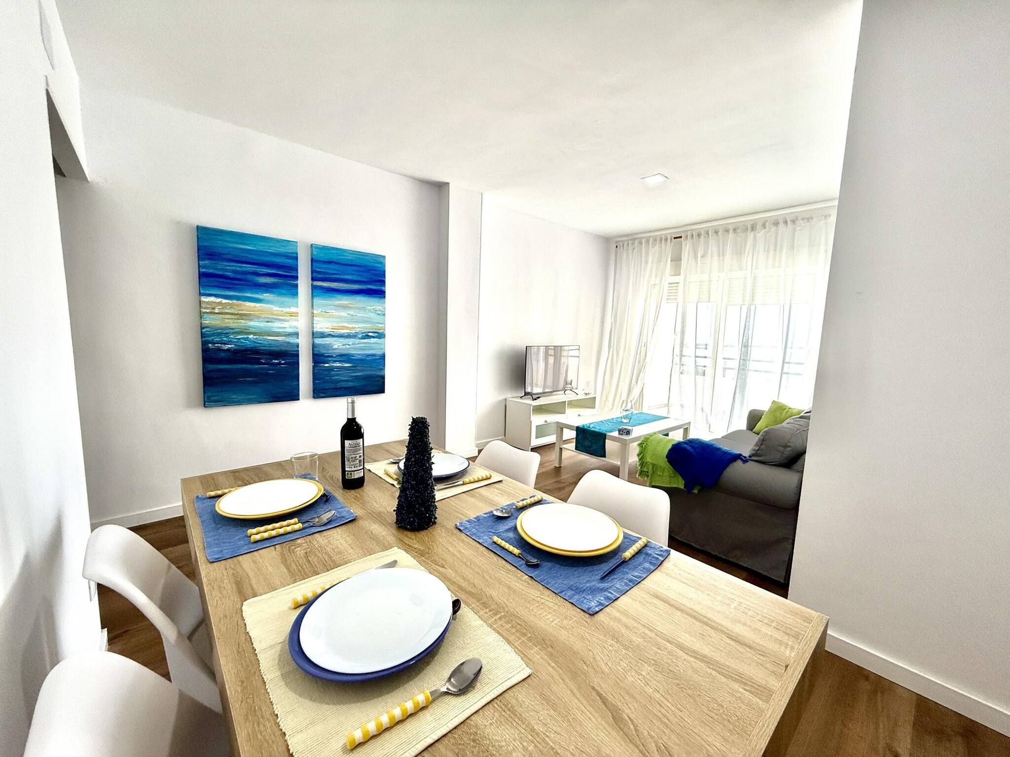 A dining area with a wooden table set for four, a TV in the background, and a living area with a sofa and a window.