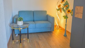 Un salon avec un canapé bleu, une petite table ronde surmontée d’un pot blanc et un grand lampadaire décoré de tournesols.