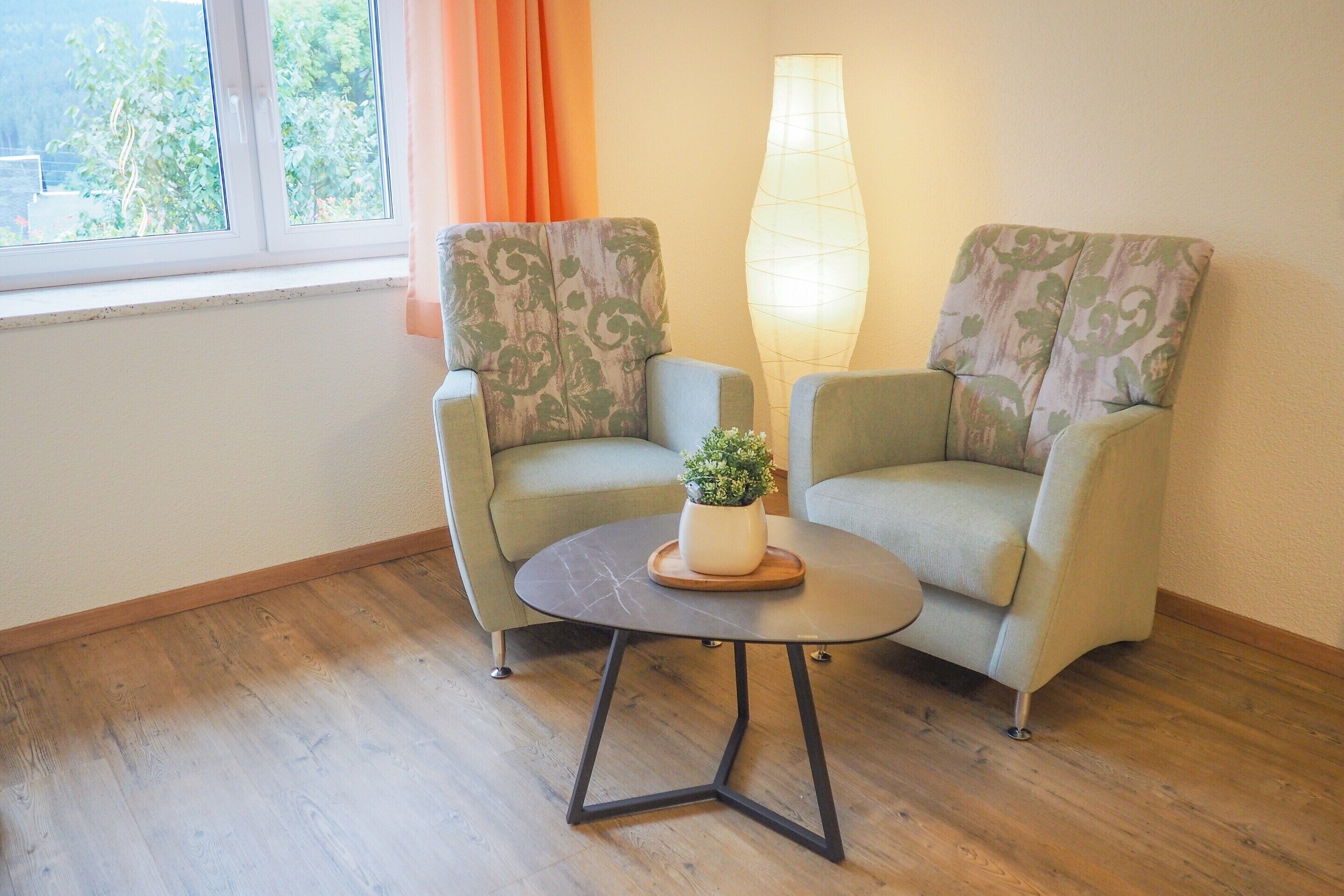 Two armchairs with a small table and a floor lamp in a room with wooden flooring and a window with curtains.