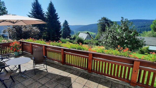 Une terrasse avec une table et des chaises, une balustrade en bois et une vue sur une vallée boisée.
