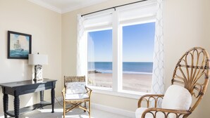 Una stanza con una grande finestra vista spiaggia, una sedia in vimini, un tavolino con una lampada e un quadro appeso al muro.