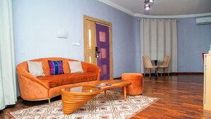 A living room with a sofa, ottoman, and coffee table, wooden flooring, and a door with a purple panel. - Eklôfè Bungalows Hôtel  (Yamoussoukro)