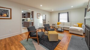 Un salon moderne avec un canapé, deux fauteuils, une table basse et un tapis. On peut apercevoir une cuisine à travers une porte ouverte.