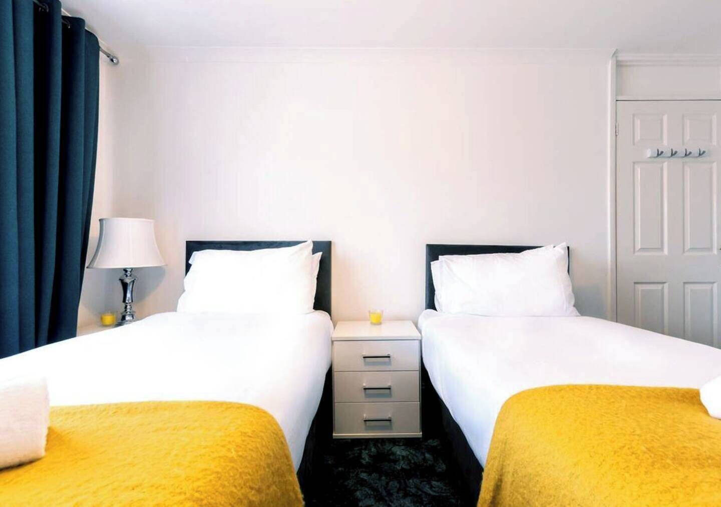 Quarto de hotel com duas camas de solteiro, mesa de cabeceira com luminária, pequena mesa com um copo e porta com cabideiro.