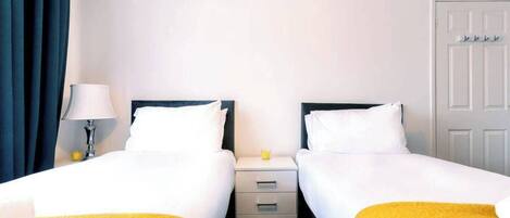 Quarto de hotel com duas camas de solteiro, mesa de cabeceira com luminária, pequena mesa com um copo e porta com cabideiro.