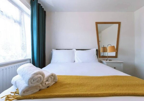 A hotel room with a bed, a window with curtains, a bedside table with a lamp, and a mirror on the wall. - Reading - 3-bed House - Sleeps 6 - Garden - Wifi (Reading)