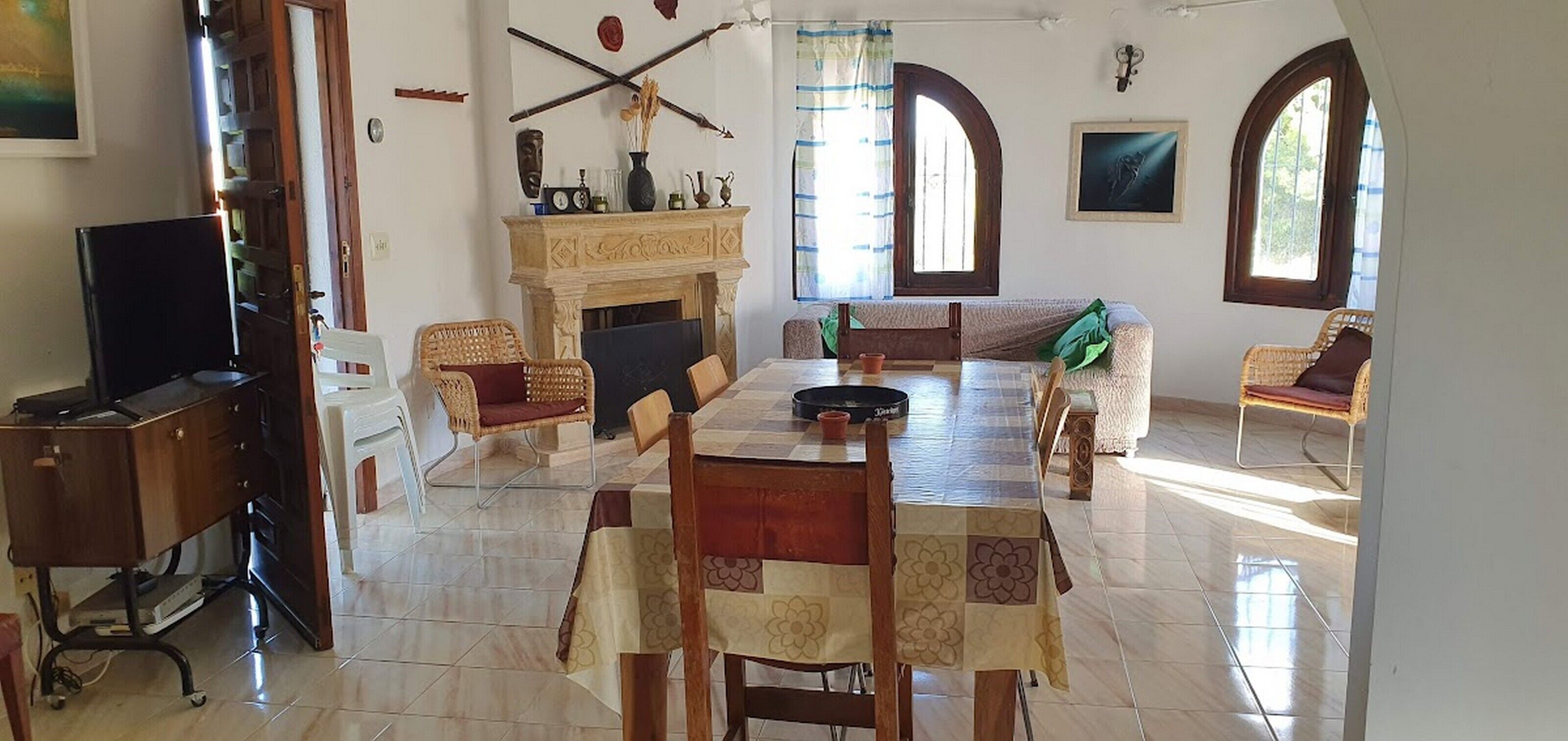 Uma sala de jantar com uma mesa posta para quatro pessoas, cercada por cadeiras de vime. Um lareira com manto aparece ao fundo, e há duas janelas com arcos.