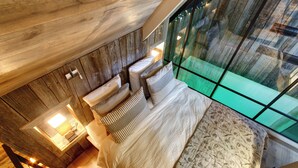 Une chambre avec un plafond en bois, un grand lit recouvert d’une literie à motifs et une vue dégagée grâce à un mur en verre.