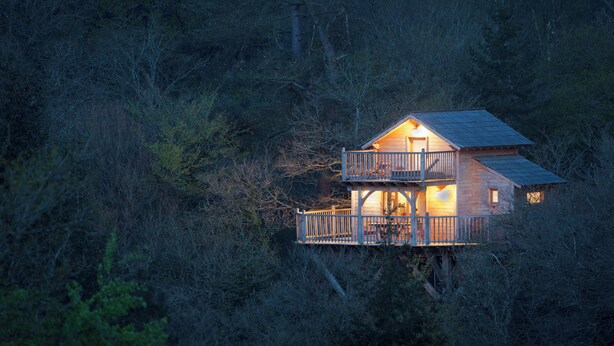 Une cabane en bois à deux étages, dotée d’un balcon et entourée d’une forêt dense.