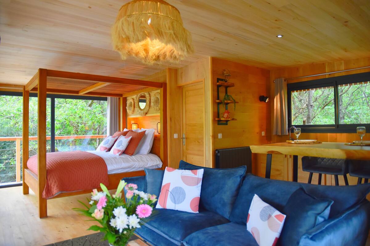 Interior de una cabaña de madera con cama, sofá, zona de comedor y un amplio ventanal con vistas al exterior.