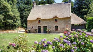 Une maison en pierre avec un toit de chaume, entourée d’un jardin bien entretenu où poussent des fleurs violettes.