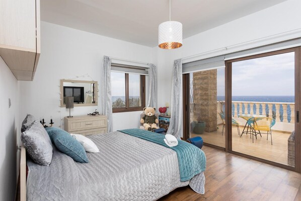 A bedroom with a large bed, a dresser, a mirror, a teddy bear, and a view of the terrace through a sliding glass door.
