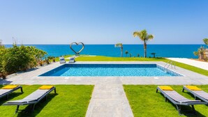 Une piscine agrémentée d’une sculpture en forme de cœur, entourée de chaises longues et de palmiers, et offrant une vue imprenable sur une mer d’un bleu limpide.
