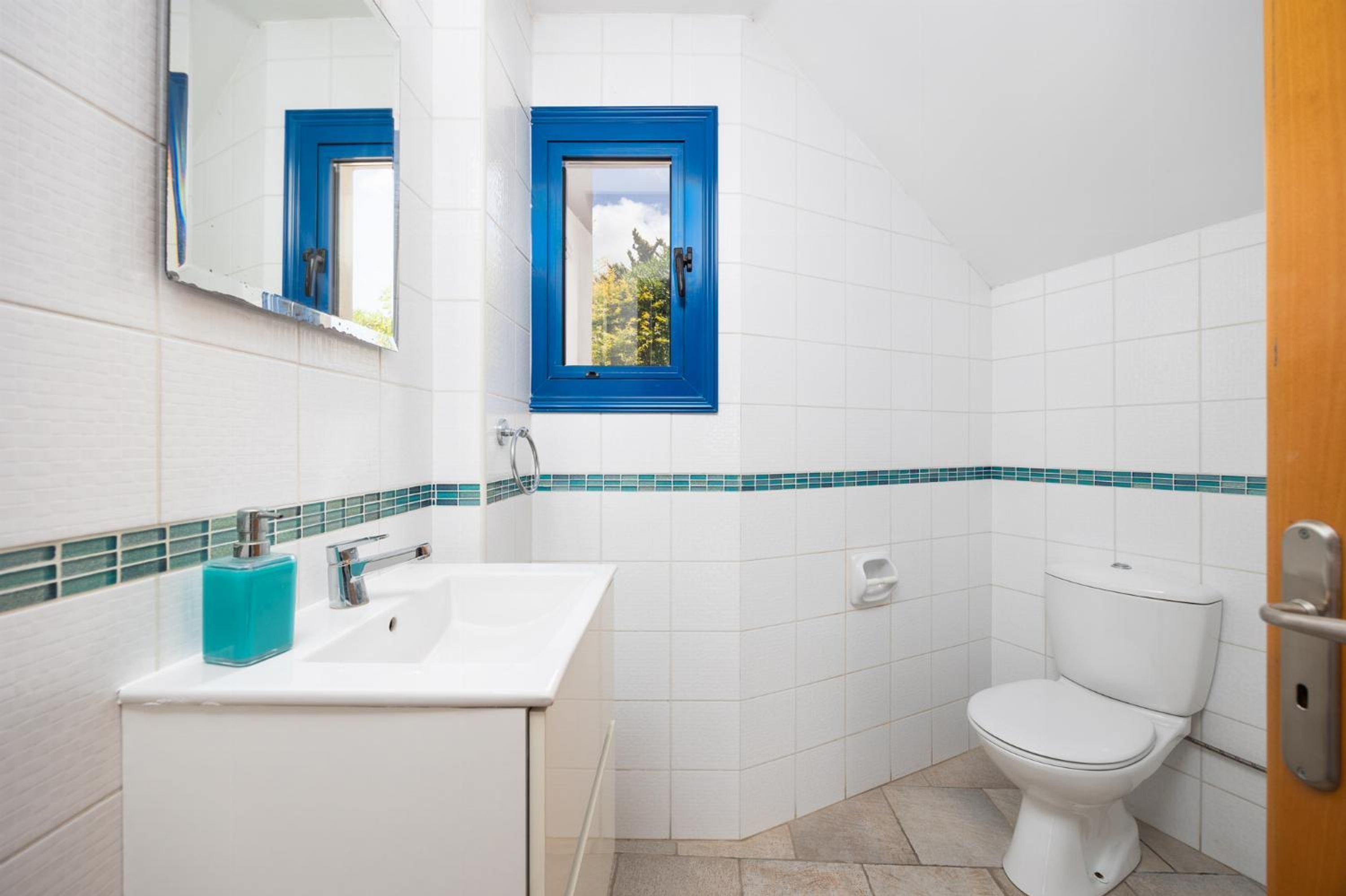 A bathroom with white tiles, a blue-framed window, a white sink, and a white toilet.
