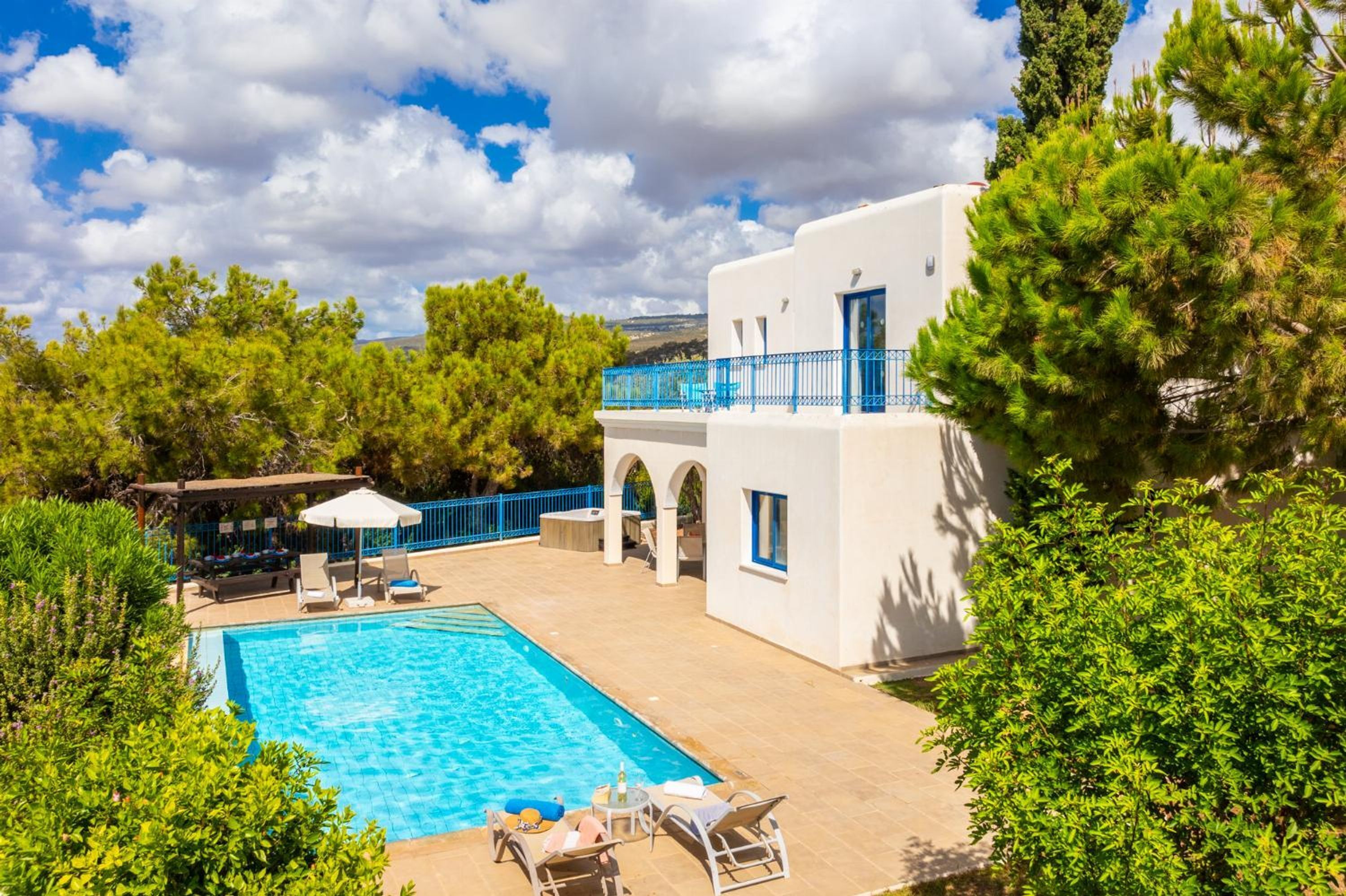 A swimming pool with lounge chairs and a pergola, surrounded by greenery and a white building with blue accents.