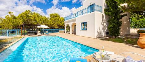 A swimming pool with lounge chairs and umbrellas, a white building with blue accents, and a pergola with a white umbrella.