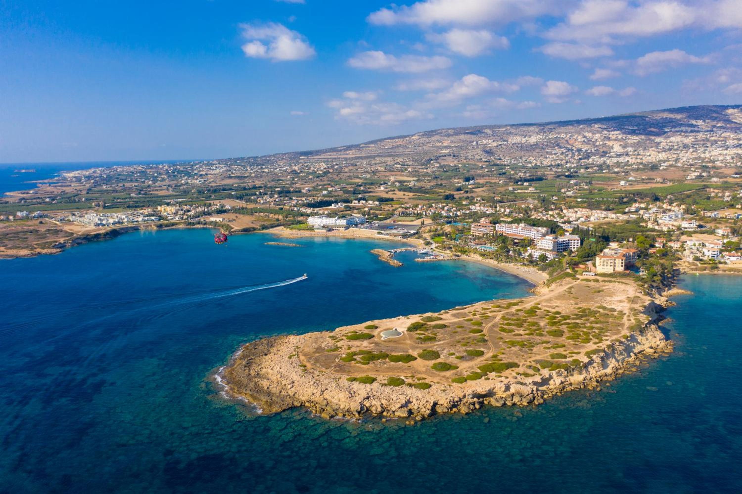 An aerial view of a coastal town with clear blue waters, a rocky peninsula, and buildings along the shore.