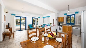 A spacious dining area with a wooden table set for six, a kitchen with modern appliances, and a view of the pool through the glass doors. - Villa Ermis in Peyia (Pegeia)