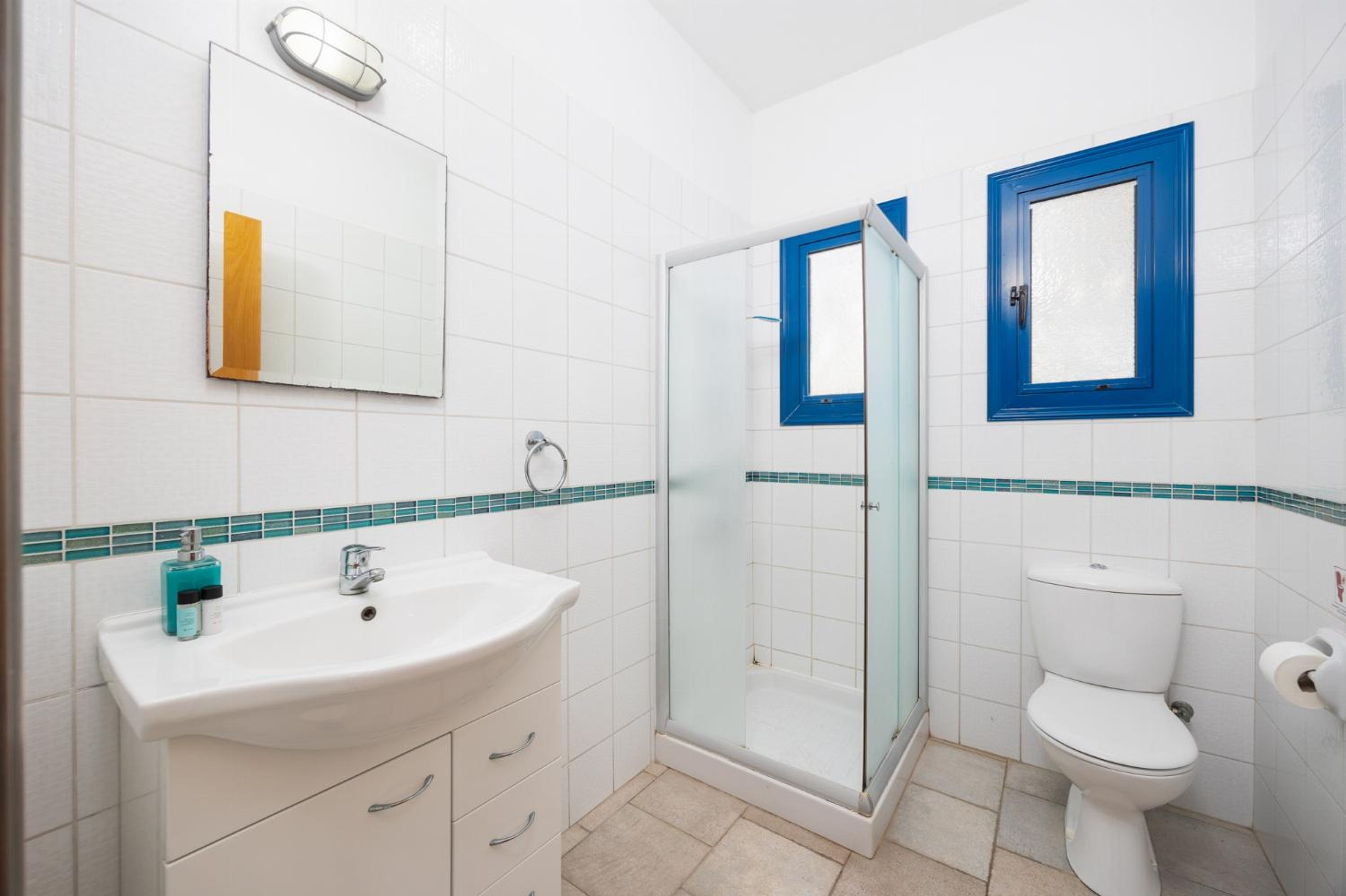 Un baño con lavamanos, espejo y retrete blancos, cabina de ducha de vidrio y ventanas con marco azul.