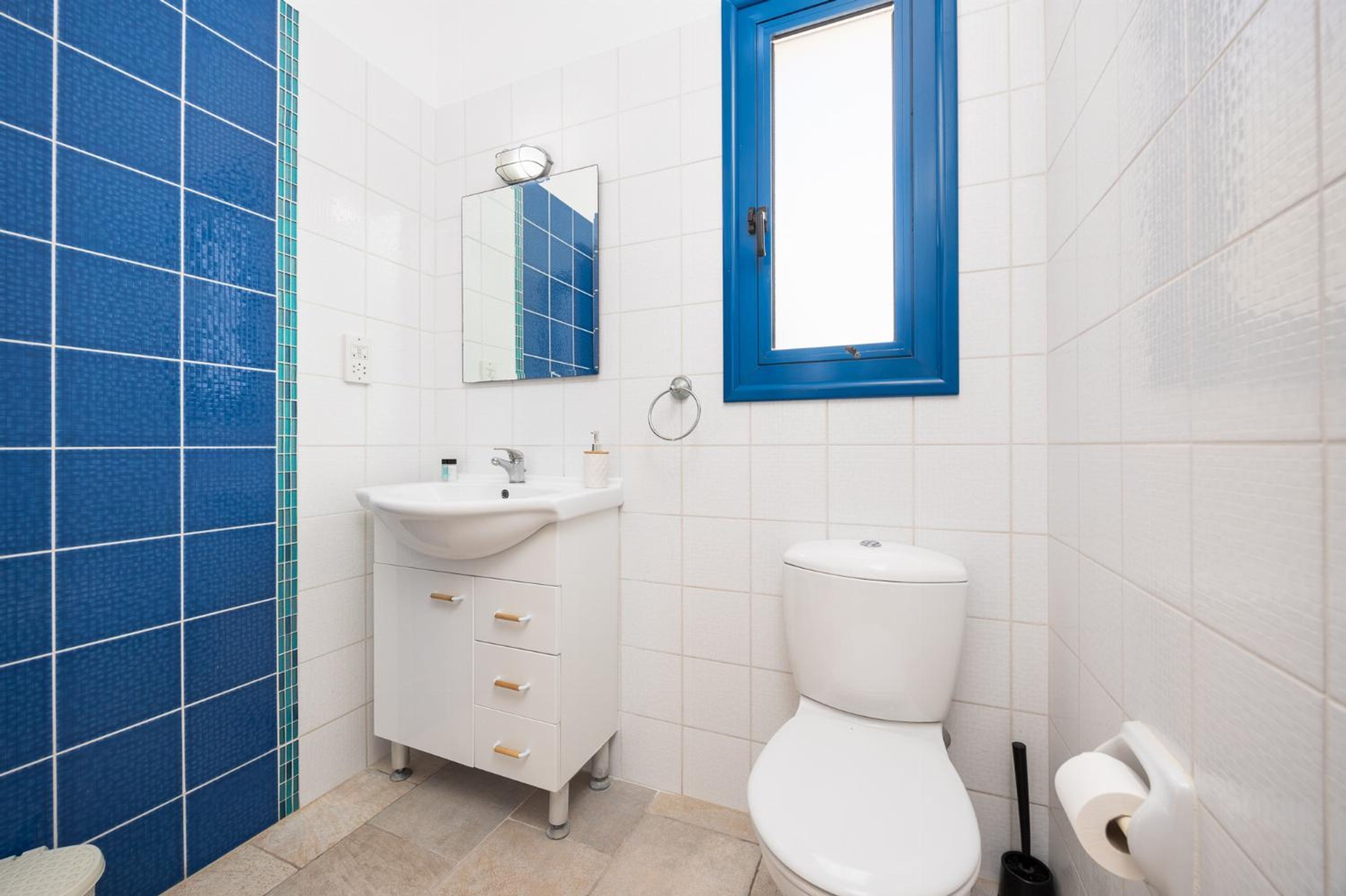 A bathroom with a white sink, mirror, and toilet, blue-tiled walls, and a blue-framed window.