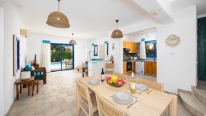 Un comedor espacioso con una mesa de madera preparada para cuatro personas, una cocina al fondo y vistas al exterior a través de un ventanal.