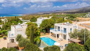 Un complejo turístico costero con edificios blancos, una piscina y vistas al mar y a las colinas.