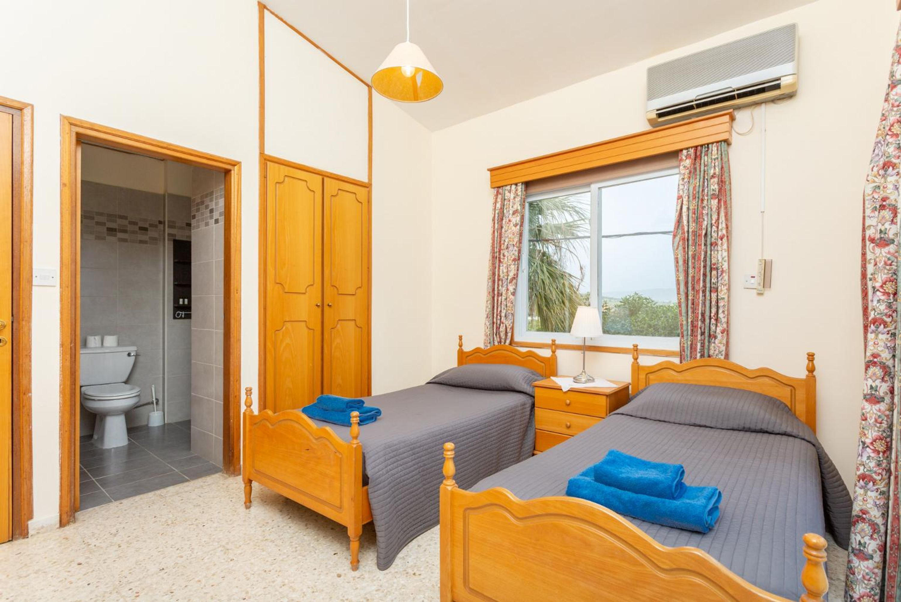 A bedroom with two wooden beds, a window with curtains, an air conditioning unit, and a bathroom visible through an open door.