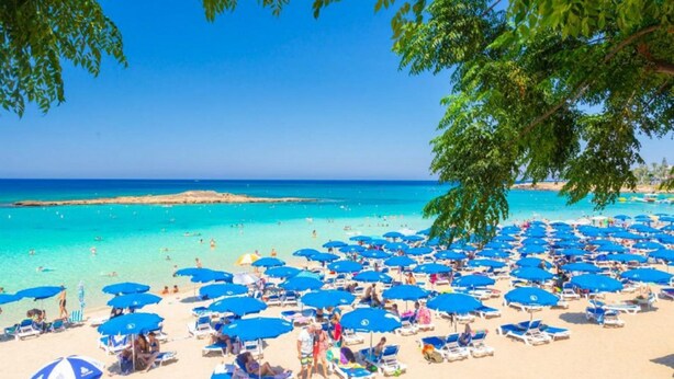 Ein Strand mit zahlreichen blauen Sonnenschirmen und Liegestühlen unter Bäumen.