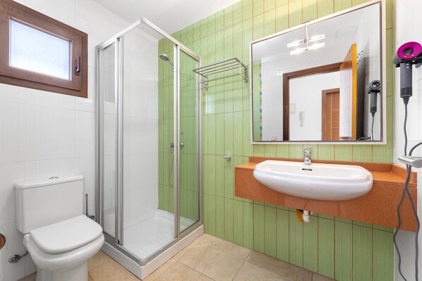 Une salle de bain comprenant des toilettes, une douche fermée par une paroi de verre, un lavabo avec un miroir au-dessus et un sèche-cheveux posé sur le comptoir.