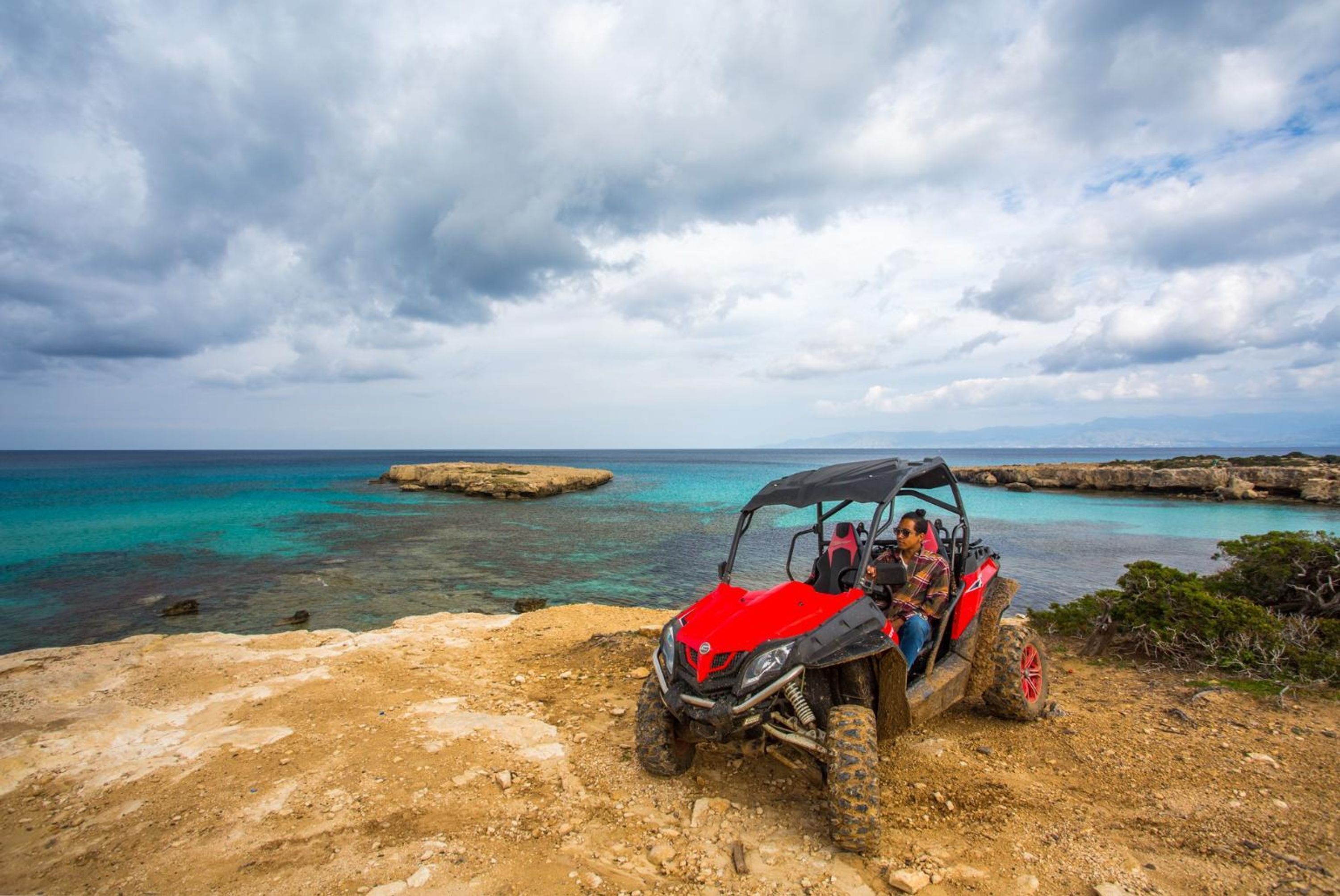 Un véhicule tout-terrain rouge est garé sur un terrain rocheux, offrant une vue sur la mer et un ciel nuageux.