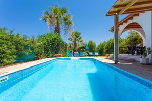 A clear blue swimming pool with a wooden pergola and lounge chairs on the right, surrounded by lush greenery and palm trees. - Villa Aristotelis in Argaka (Argaka)