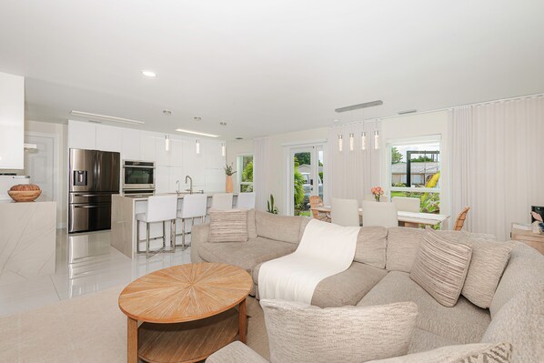 A modern living room with a sectional sofa, a wooden coffee table, and a kitchen area with stainless steel appliances. - Luxury Beach Home Salt Water Pool Kid Pet Friendly (Boca Raton)
