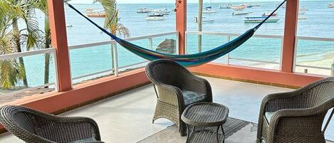 A covered outdoor seating area with wicker furniture and a hammock, overlooking a beach with boats.