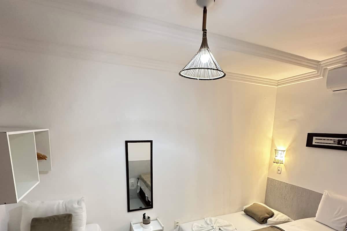 A hotel room with two beds, a small bedside table, a mirror, a hanging light fixture, and a wall-mounted air conditioning unit.