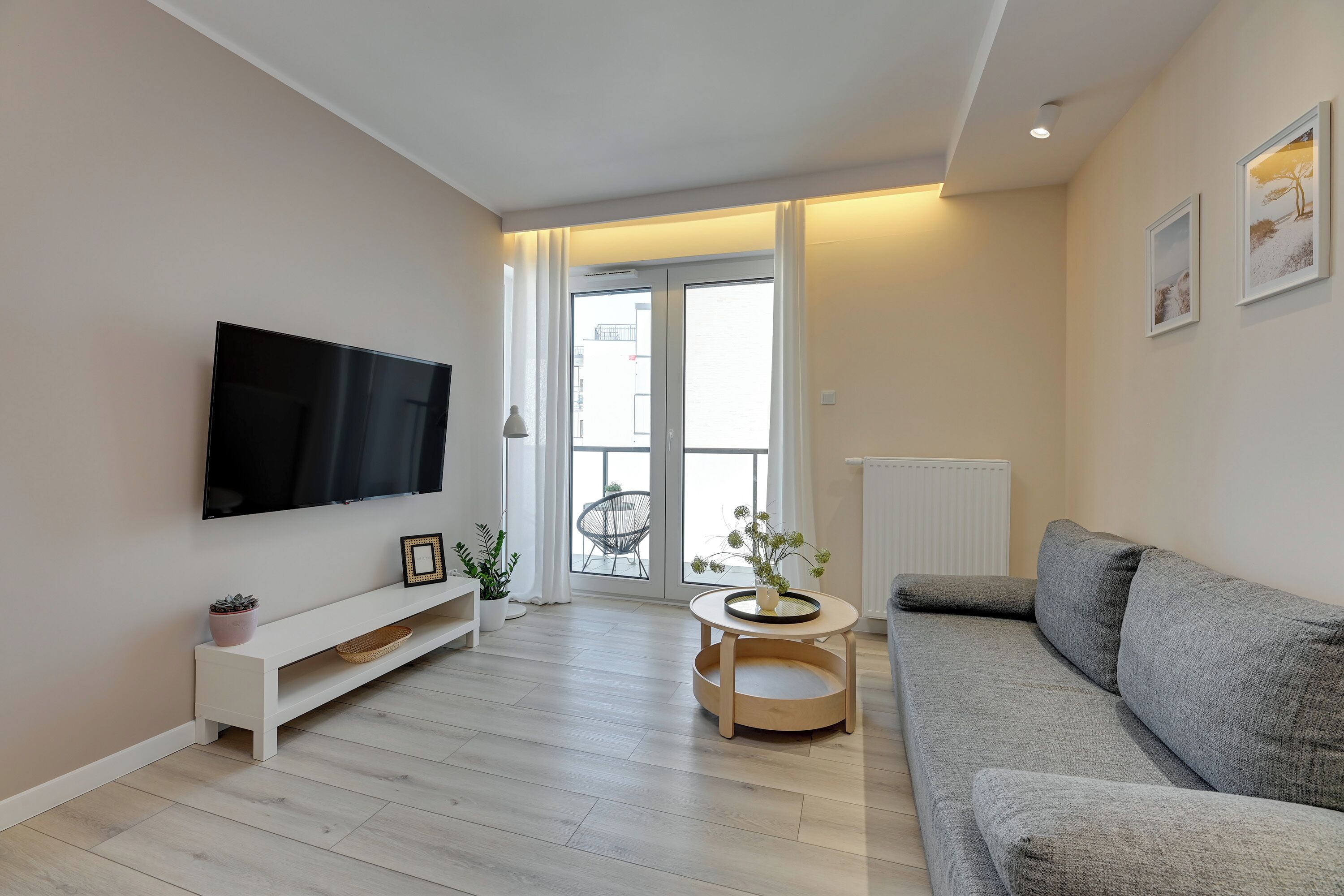 Una sala moderna con un televisor de pantalla plana, un sofá gris, una consola blanca y una pequeña mesita de madera con un florero.