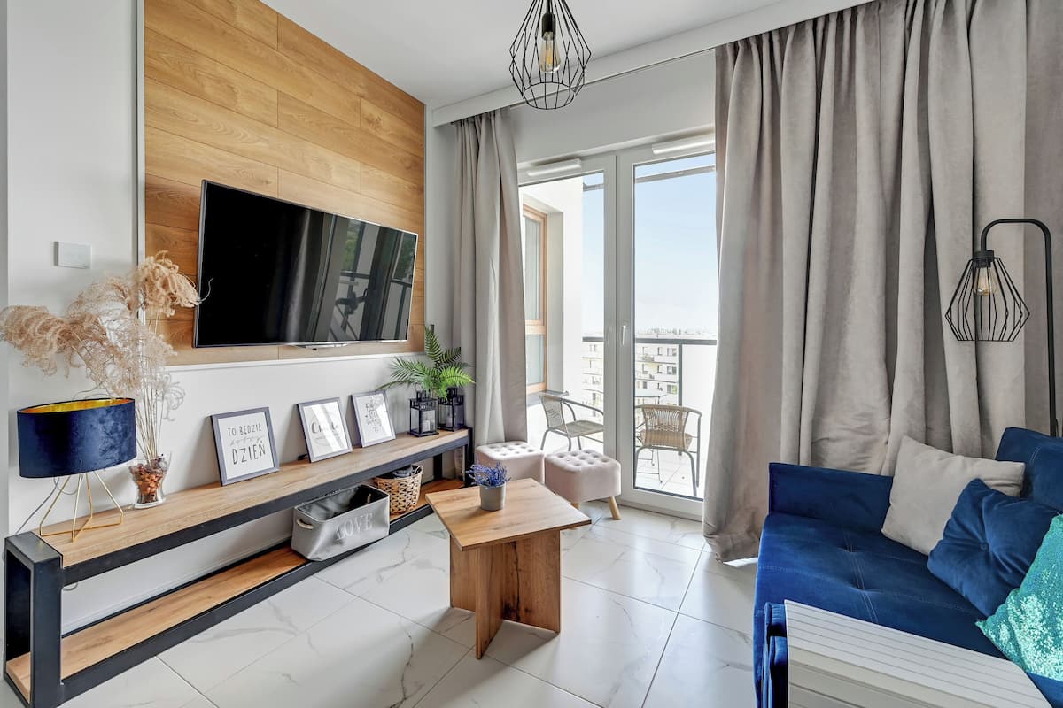 A modern living room with a flat-screen TV, wooden panel wall, a blue sofa, and a balcony with curtains.