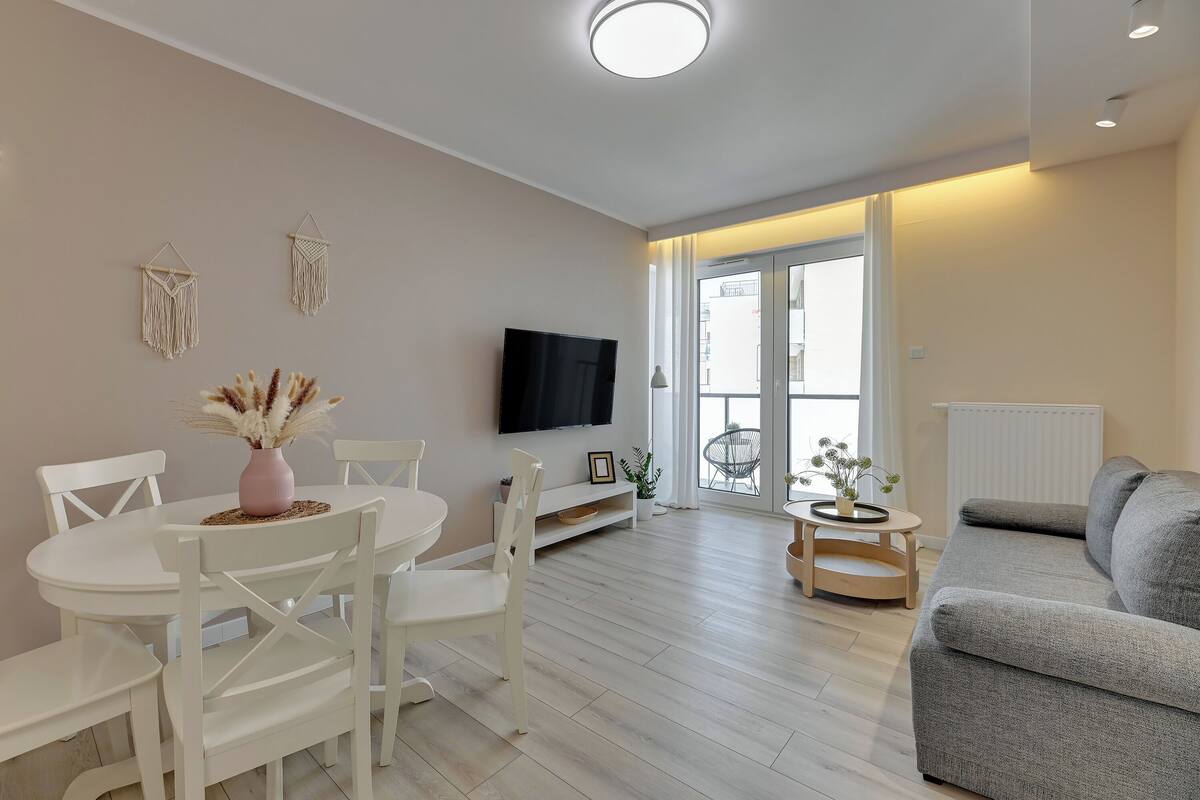 A modern living room with a grey sofa, white dining table with chairs, a flat-screen TV mounted on the wall, and a large window with a view of the city.
