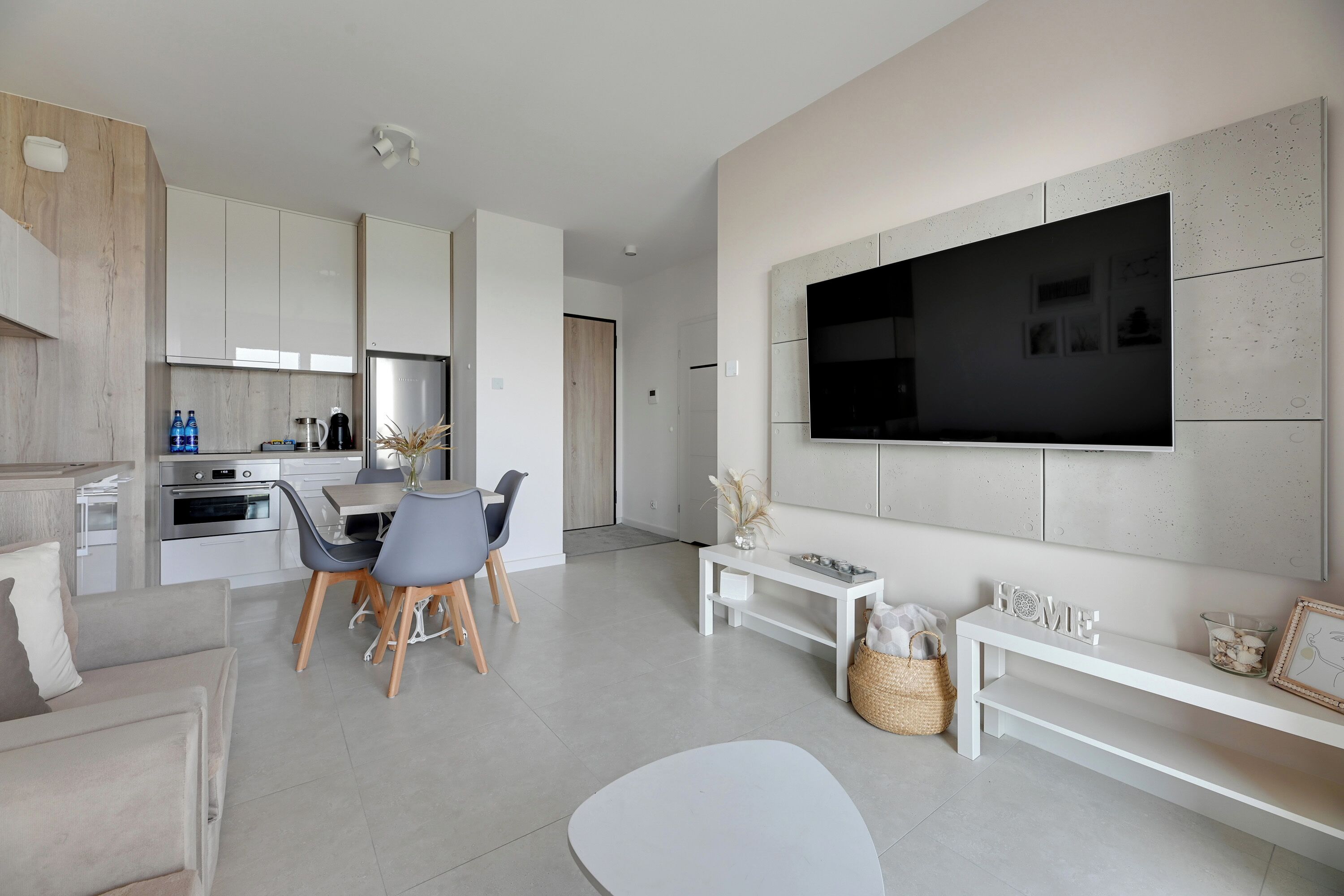 Uma sala de estar moderna com uma televisão de tela plana grande fixada na parede, uma área de jantar com mesa e cadeiras, e uma cozinha com armários e eletrodomésticos brancos.