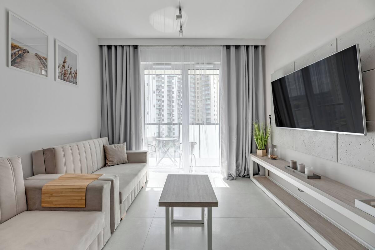 A modern living room with a sofa, a coffee table, a TV mounted on the wall, and a large window with curtains.