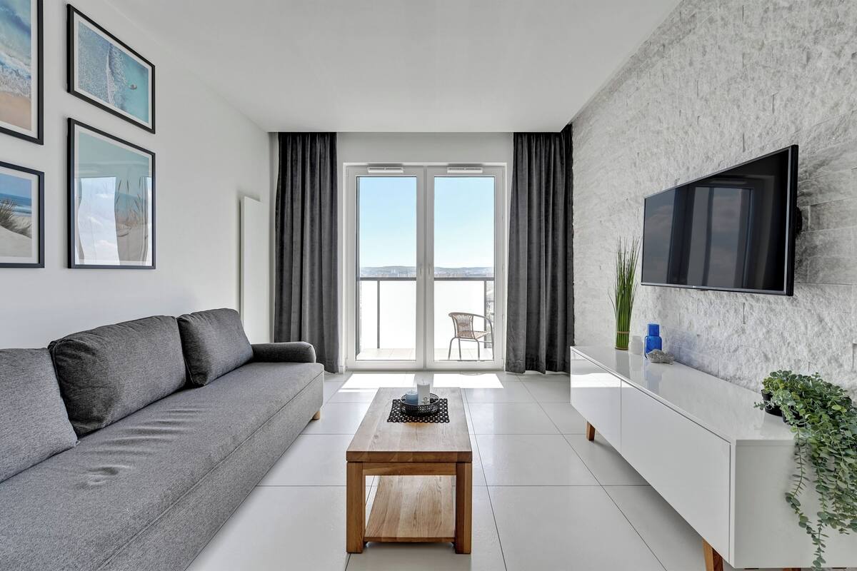 A modern living room with a grey sofa, a wooden coffee table, a white TV stand, and a large window with curtains.