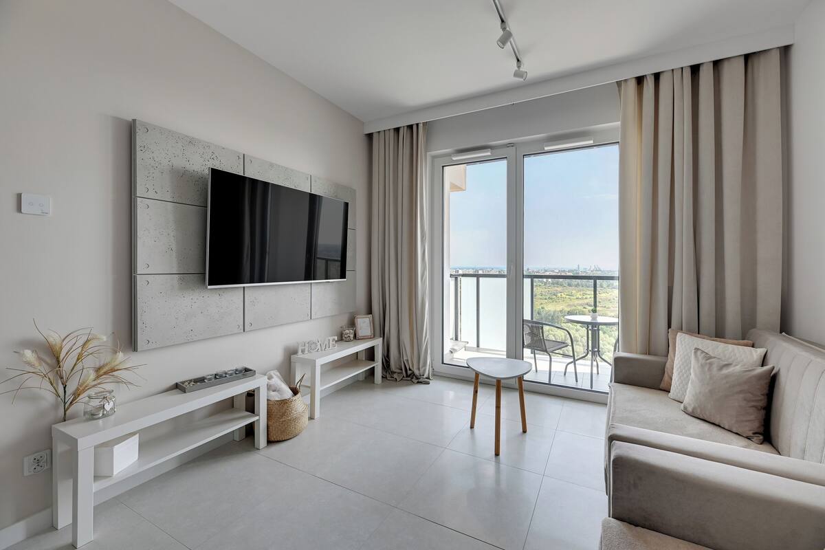 A modern living room with a large flat-screen TV, a sofa, a small table, and a balcony with a view.