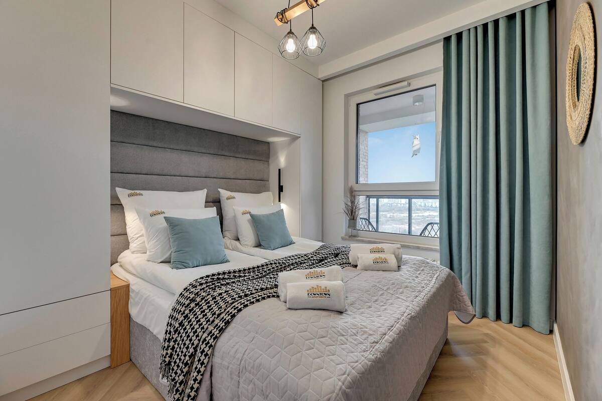 A modern bedroom with a large bed, grey headboard, white bedding, and a view of the city through a window with curtains.