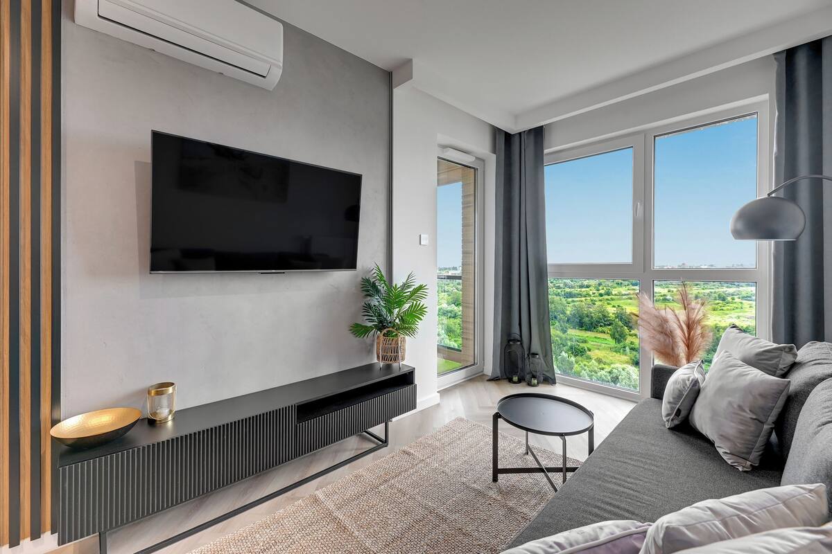 A modern living room with a flat-screen TV mounted on the wall, a black entertainment unit, a round coffee table, and a grey sofa with cushions.