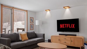 En moderne stue med en grå sofa, en rød skammel, et træskab og et fjernsyn, der viser "NETFLIX".