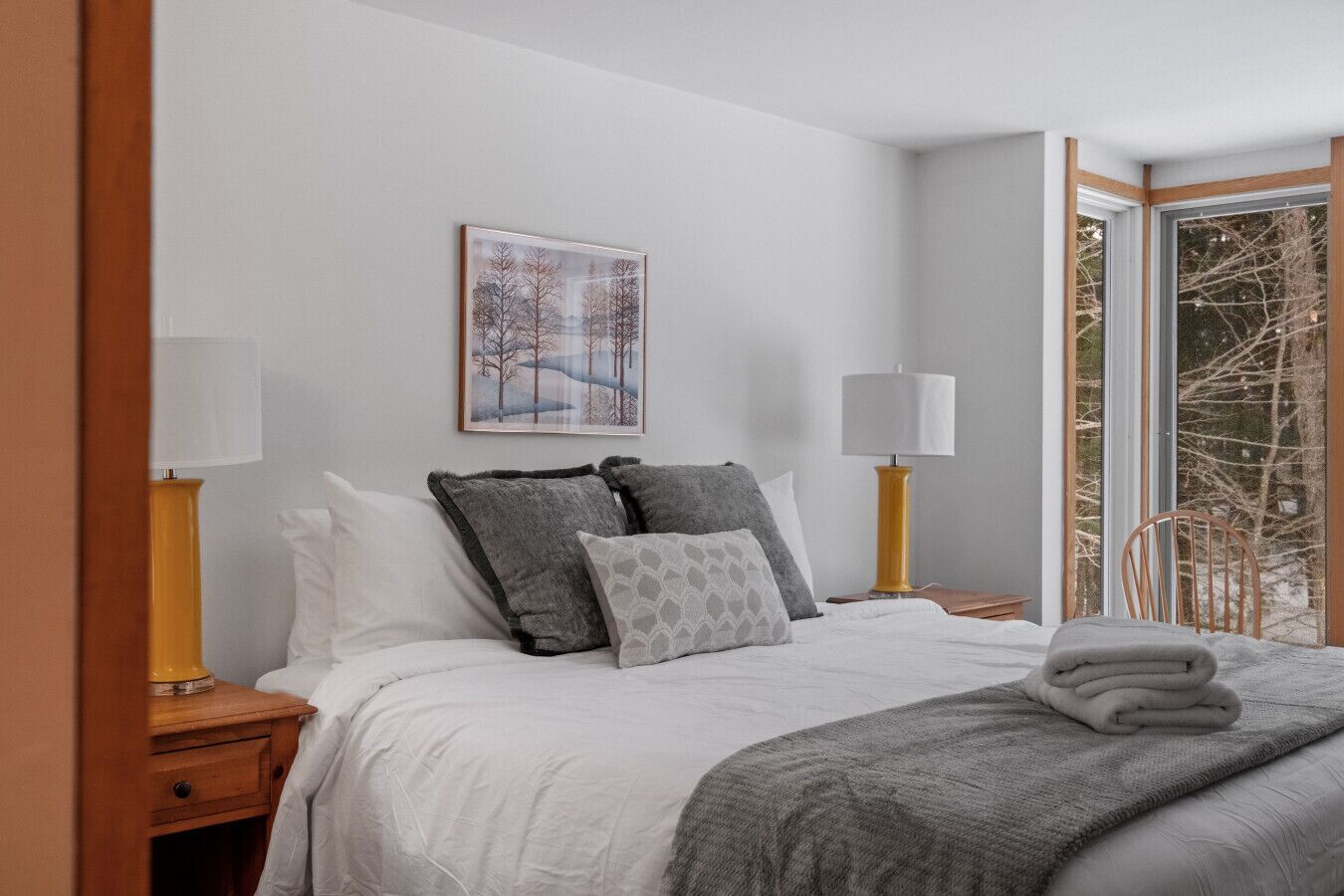 A neatly made bed with a white comforter and gray pillows, a framed picture on the wall, and a window with a view of bare trees.
