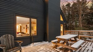 En træterrasse med et picnicbord og stole, en træstol og et vindue der kigger ud over et rum med en seng og en lampe.