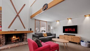 En moderne stue med pejs, en rød sofa, en grå sofa, et træ-TV-bord og et fladskærms-TV der viser "NETFLIX".