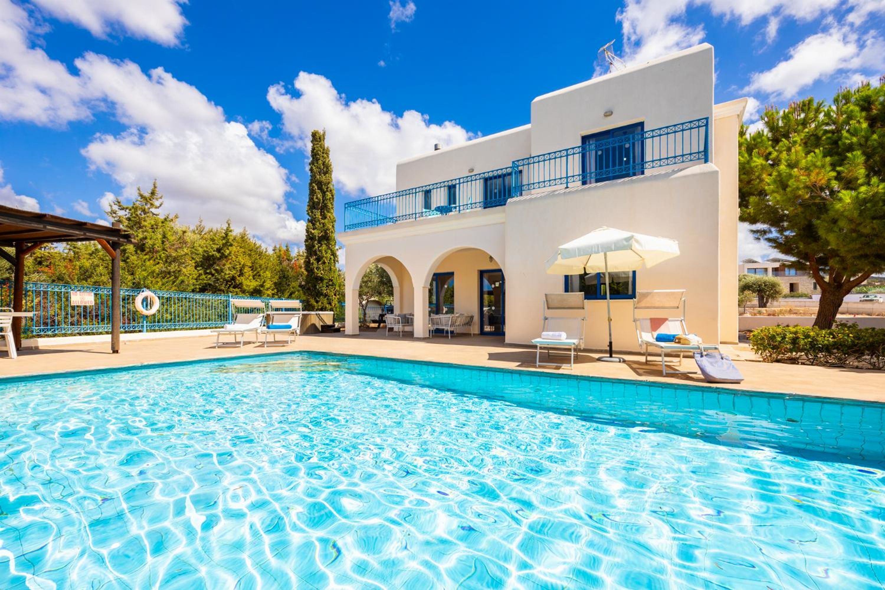 Une piscine avec des chaises longues et des parasols, un bâtiment blanc agrémenté de touches de bleu, et un ciel d’un bleu limpide.