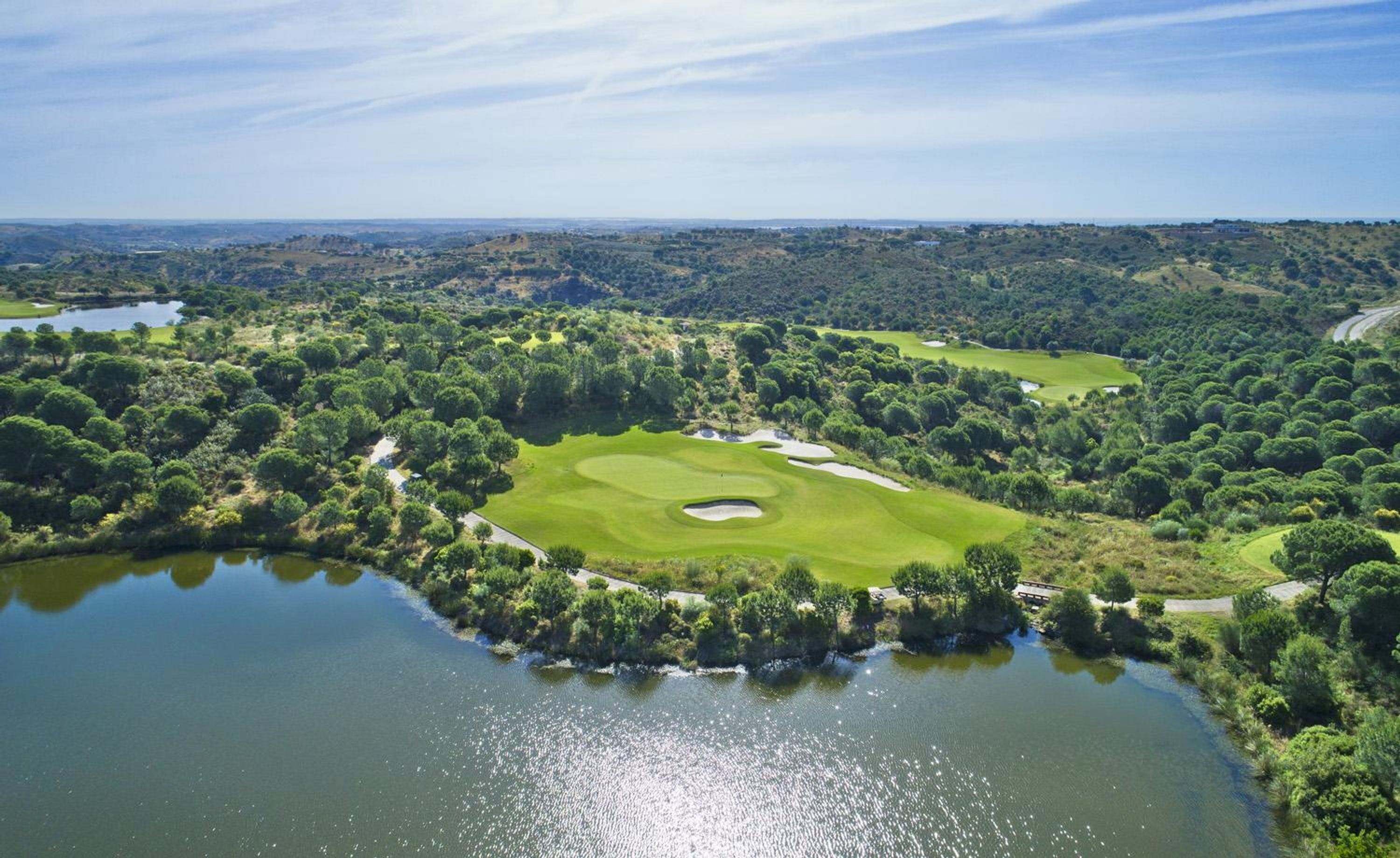 Un campo de golf con un estanque, rodeado de árboles densos y un paisaje ondulado.